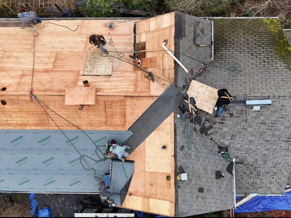 Aerial view of workers replacing a house roof; half of the roof shows new plywood, while the other half has old gray shingles. Ropes and equipment are visible, with workers actively installing materials.