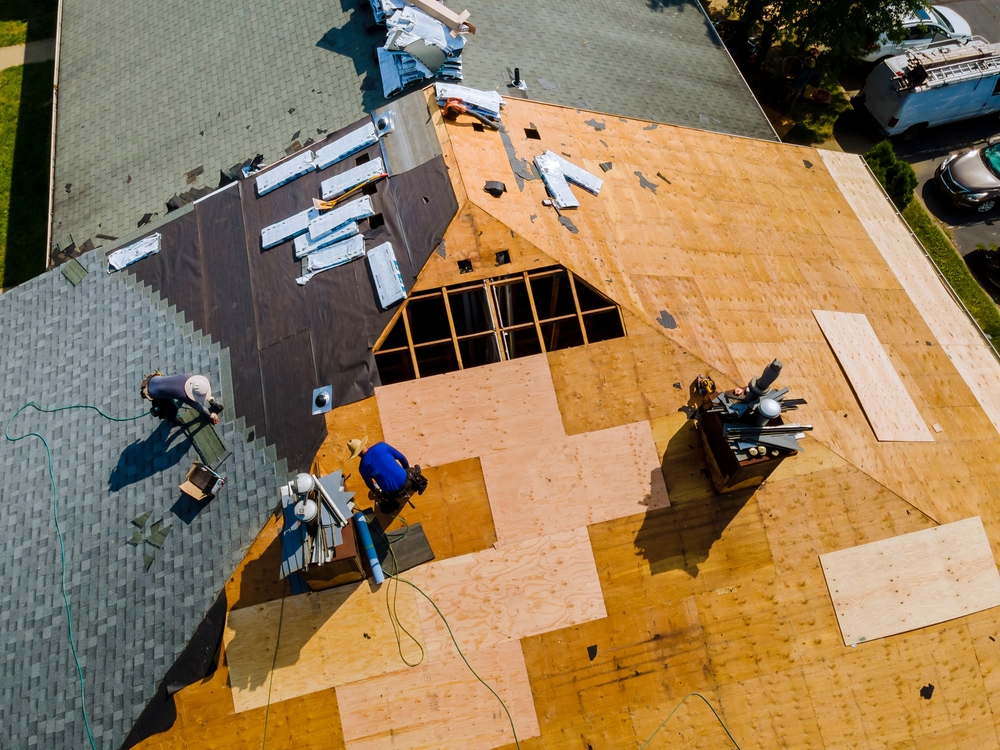 Aerial view of a house roof under renovation. Workers are installing new plywood and shingles. Some areas are covered with black roofing felt. Tools and equipment are scattered on the roof. Partially completed sections show progress.