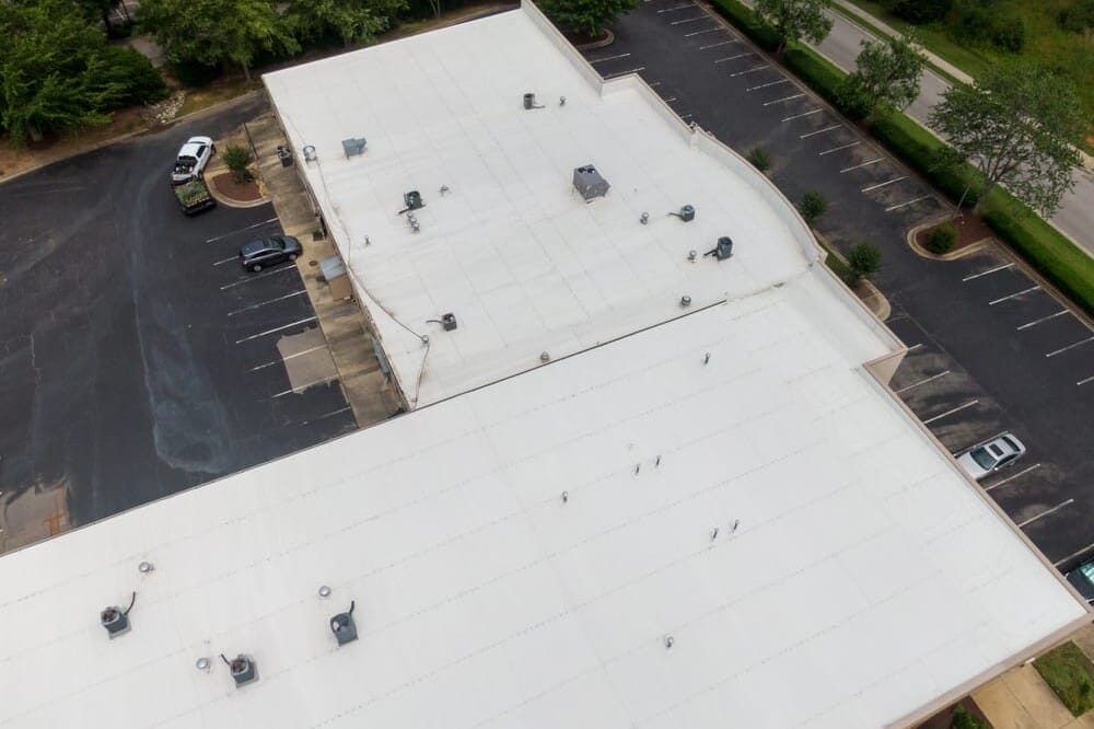 Aerial view of a large, white-roofed commercial building surrounded by mostly empty parking lots. Trees line the edges of the parking area, and a few cars are parked. The roof features multiple HVAC units and vents.