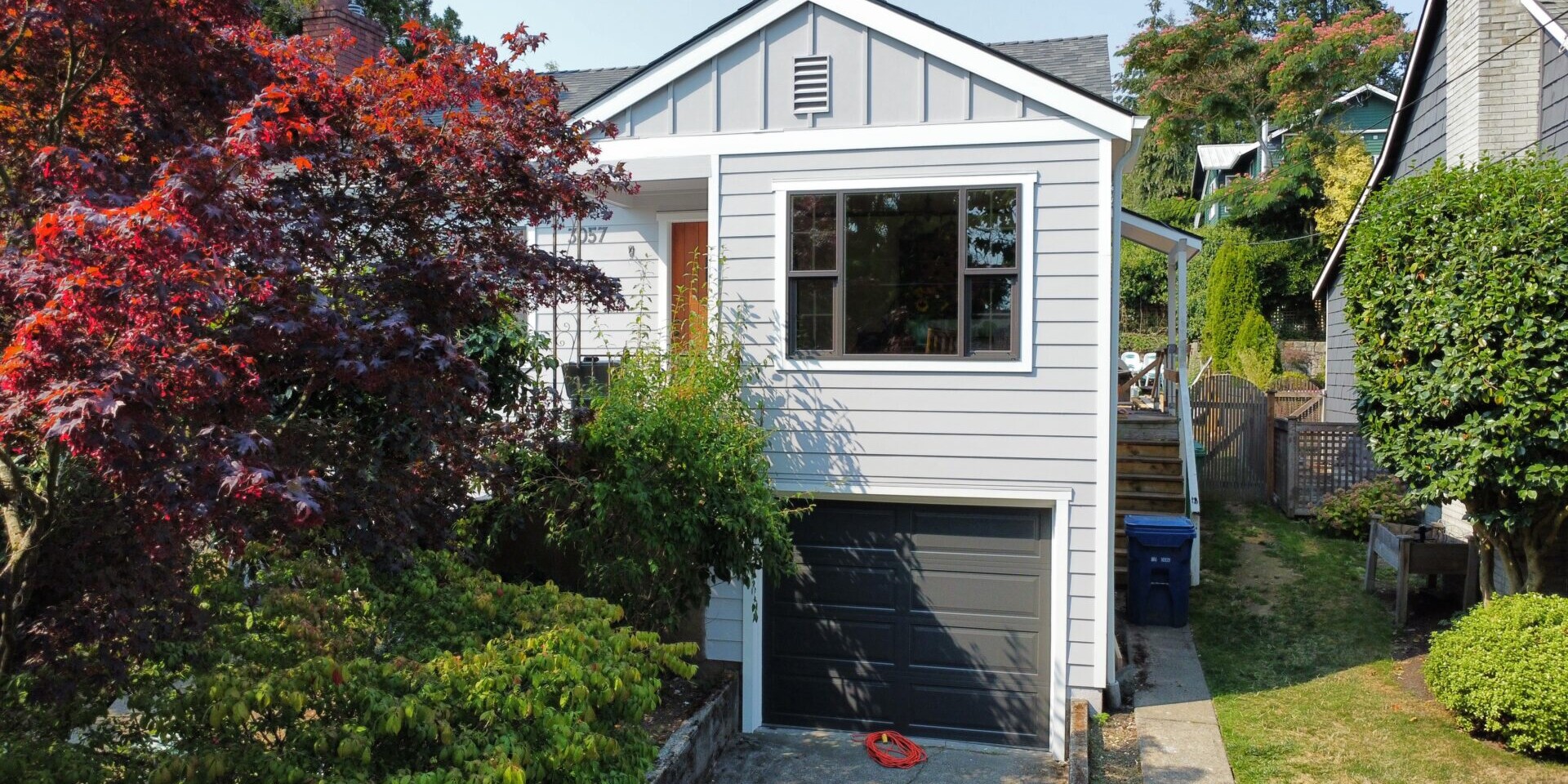 A small, white house with a dark garage door sits between trees. The yard has a red-leafed tree and green shrubs. A driveway leads to the garage, and there are steps to the side of the house. A blue recycling bin is visible.