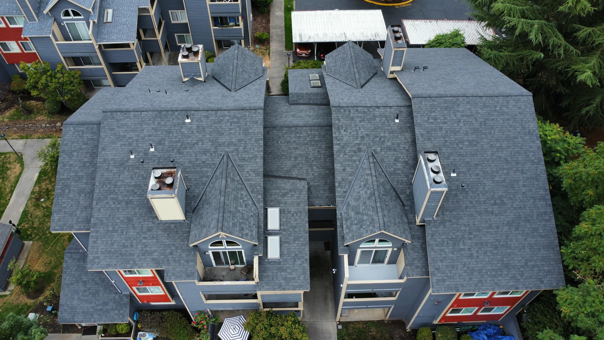 Aerial view of a modern apartment complex with gray rooftops and multiple chimneys. The buildings have red and beige accents. Surrounding the complex, there are trees and a multi-story building nearby.
