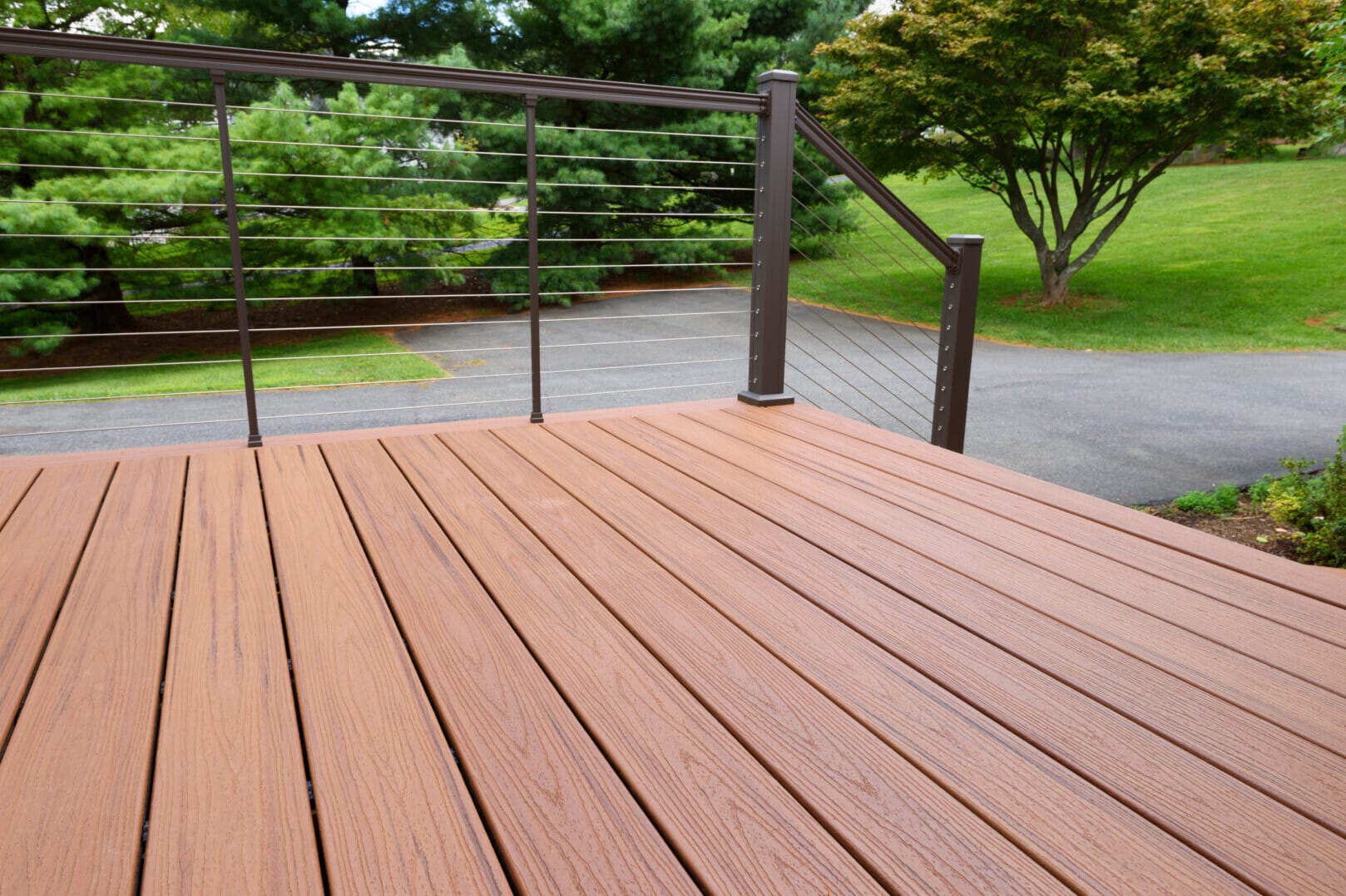A wooden deck with horizontal cable railing overlooks a paved area and a green lawn with trees in the background. The deck boards are arranged lengthwise, creating a clean and modern look.
