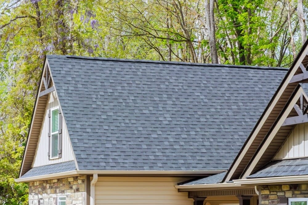 A house with a steeply pitched roof covered in dark gray shingles. The exterior features a mix of stone and siding. Surrounding the house are tall trees with green leaves.