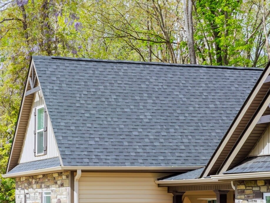 A house with a steeply pitched roof covered in dark gray shingles. The exterior features a mix of stone and siding. Surrounding the house are tall trees with green leaves.