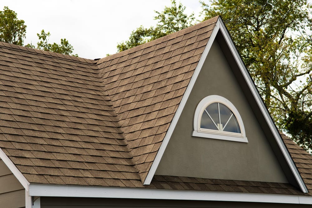 A close-up view of a house roof with brown asphalt shingles, featuring a gable with a small arched window and white trim, surrounded by green trees in the background.