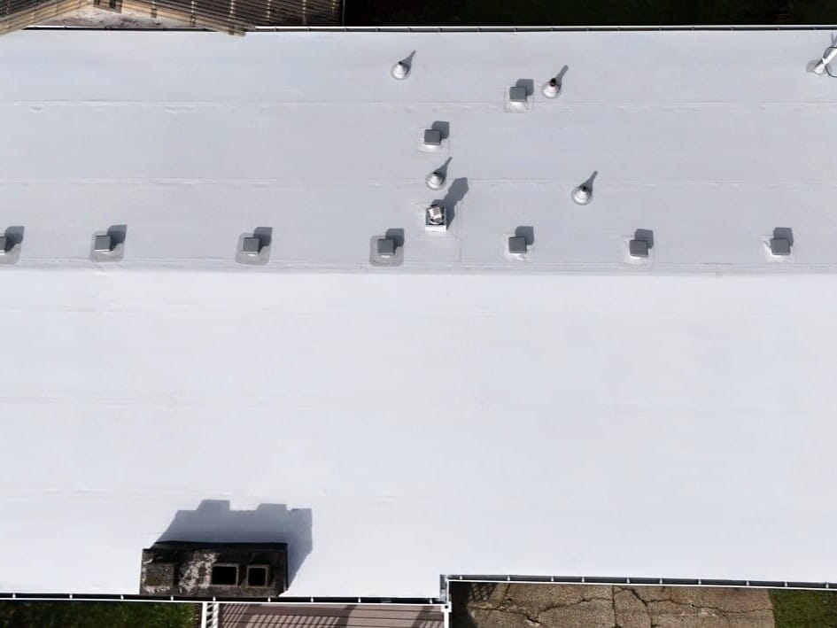 Aerial view of a large white flat roof with several vents and units evenly spaced; trees and pavement are visible at the edges.