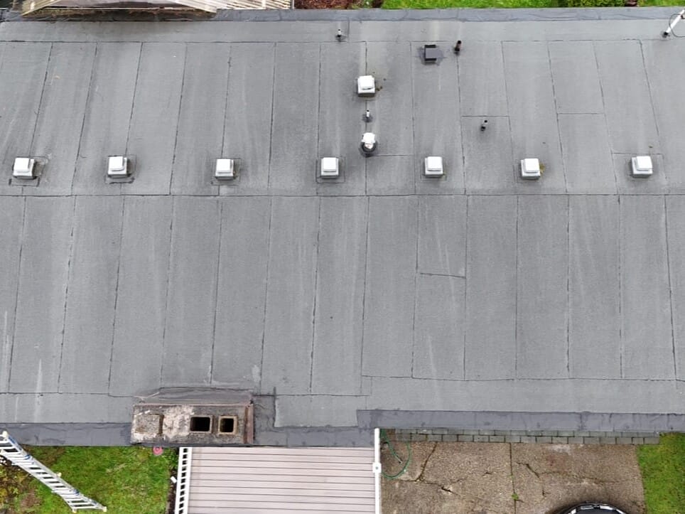 Aerial view of a flat, gray roof with multiple vents and a small damaged area near one edge. The surrounding areas feature green lawns, shrubs, and a driveway.