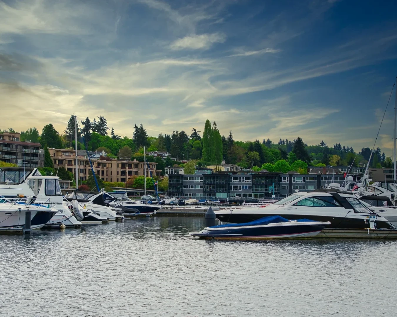 Boats and yachts docked at a marina with residential buildings and trees in the background under a partly cloudy sky at dusk.