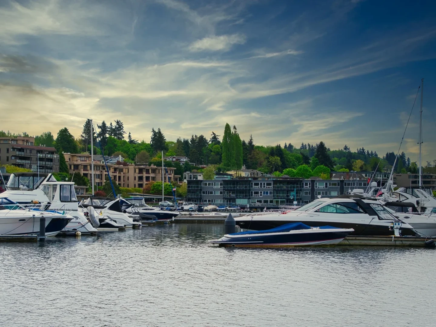 Boats and yachts docked at a marina with residential buildings and trees in the background under a partly cloudy sky at dusk.