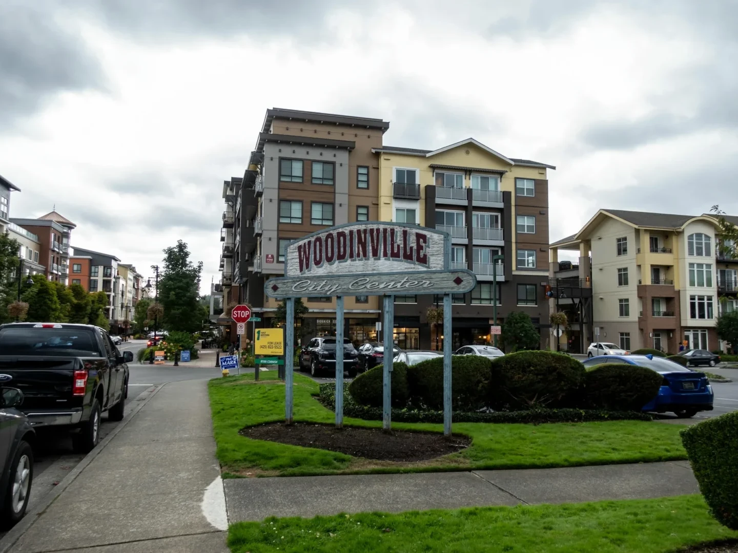 A large sign reading Woodinville City Center stands on a grassy median with apartment buildings, parked cars, and a cloudy sky in the background.