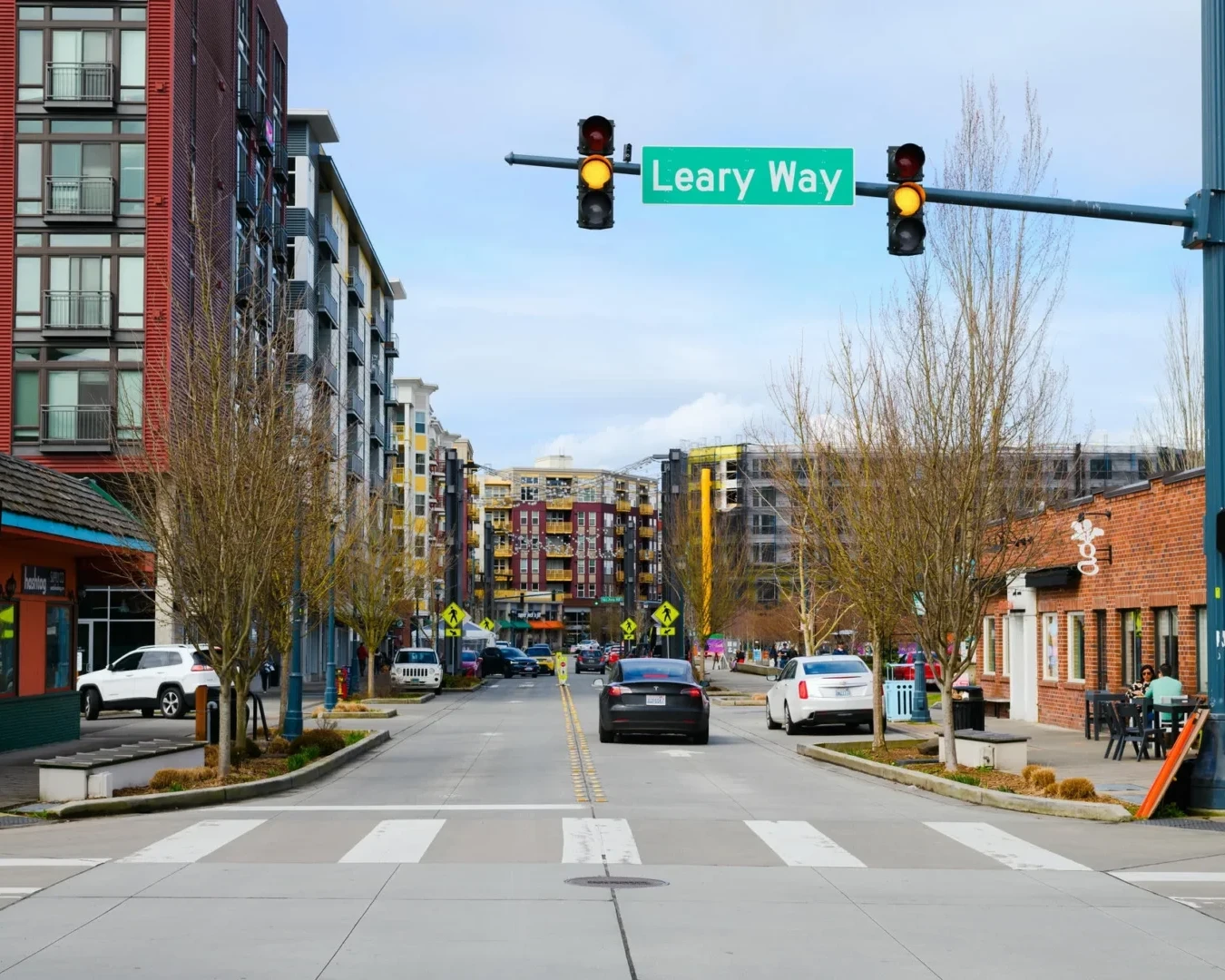 A city street scene at an intersection with the green Leary Way street sign overhead, lined with modern apartments, shops, crosswalk, pedestrians, cars, and bare trees on both sides of the road.