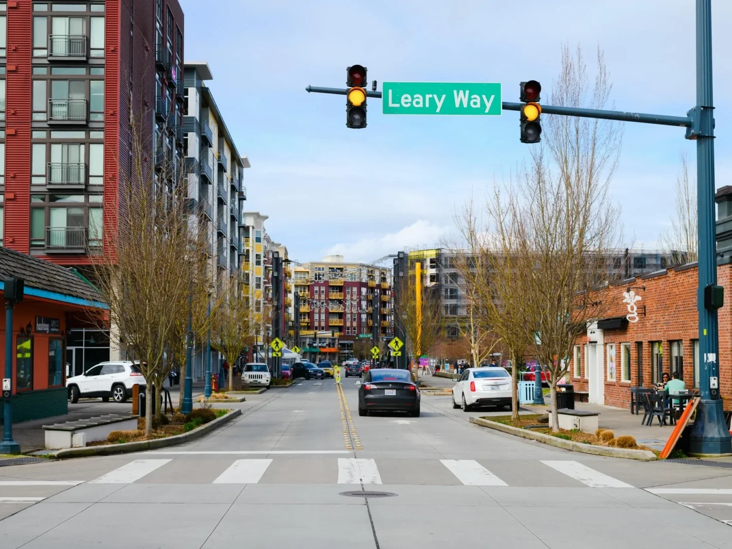 A city street scene at an intersection with the green Leary Way street sign overhead, lined with modern apartments, shops, crosswalk, pedestrians, cars, and bare trees on both sides of the road.