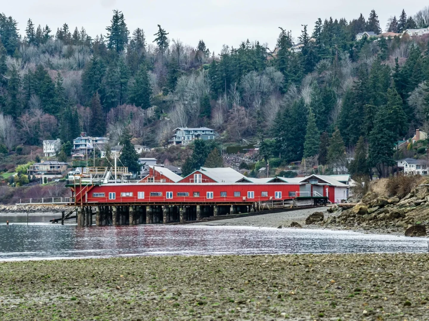A large red building on wooden stilts extends over the water near a rocky shoreline, with houses and dense evergreen trees covering the hillside in the background.