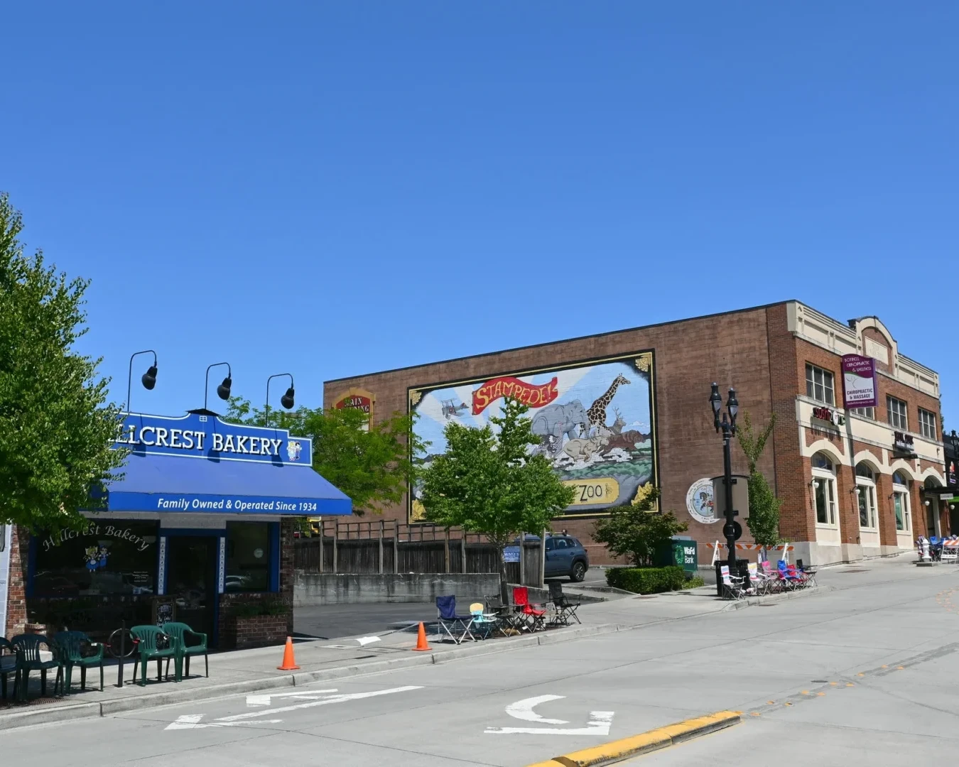 A sunny street scene shows Hillcrest Bakery with a blue awning, outdoor seating, and a large mural on a brick building. Other businesses and a few trees line the sidewalk under a clear blue sky.