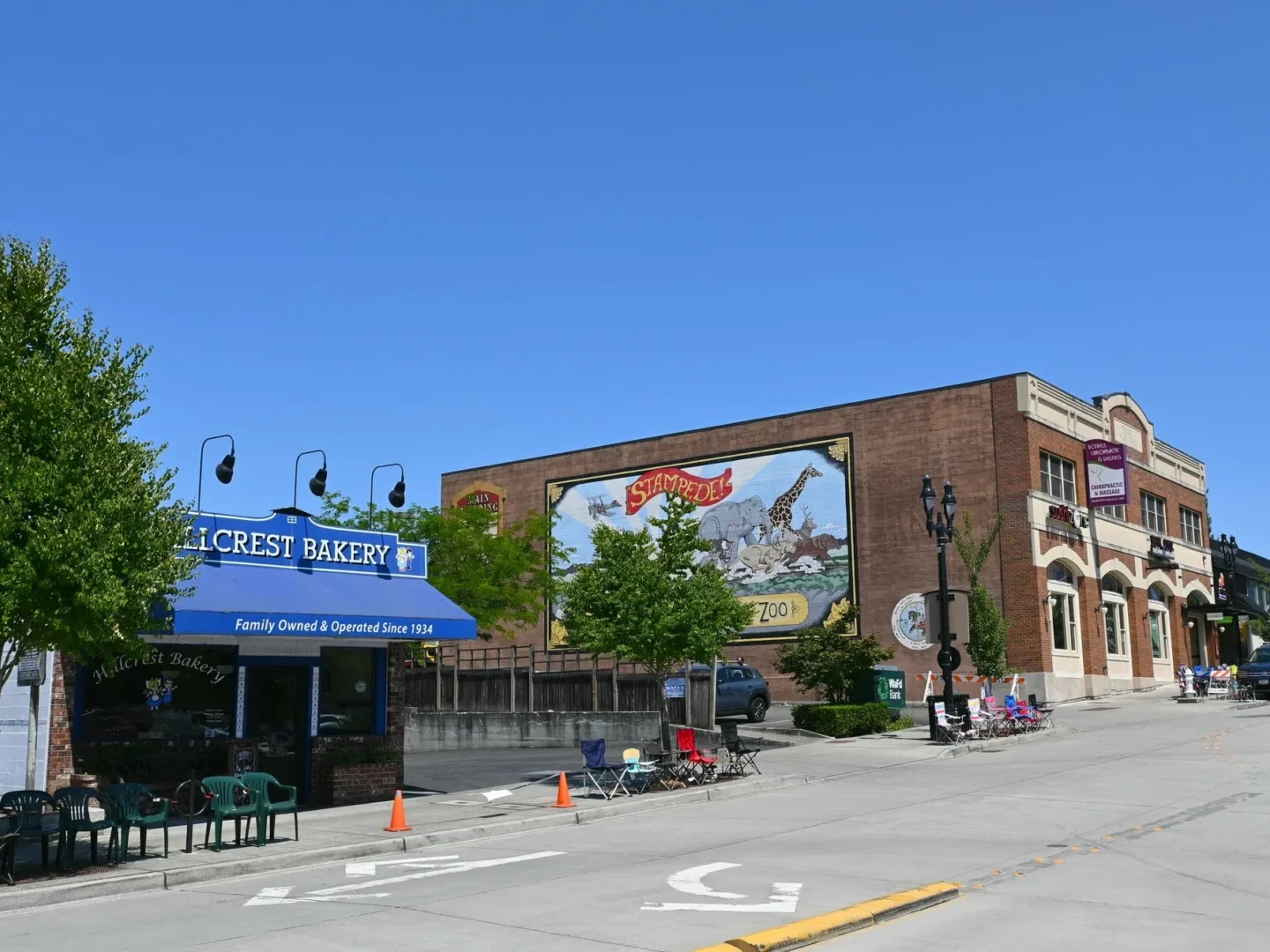 A sunny street scene shows Hillcrest Bakery with a blue awning, outdoor seating, and a large mural on a brick building. Other businesses and a few trees line the sidewalk under a clear blue sky.