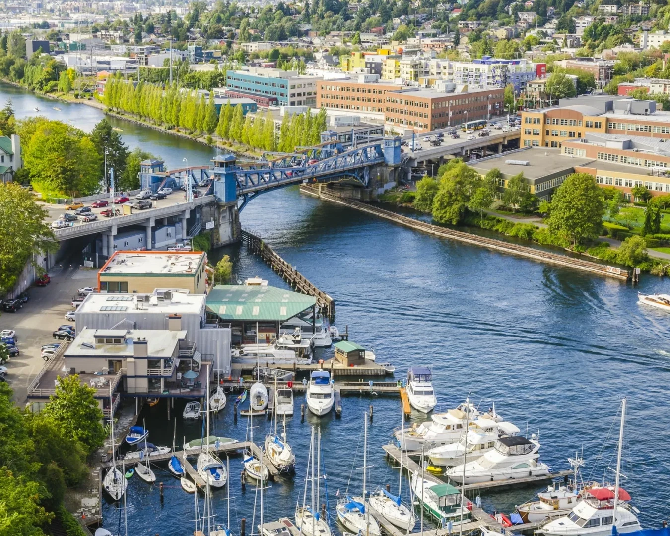 Aerial view of a marina with boats docked, a blue drawbridge over a canal, and surrounding buildings and greenery, with a cityscape in the background.