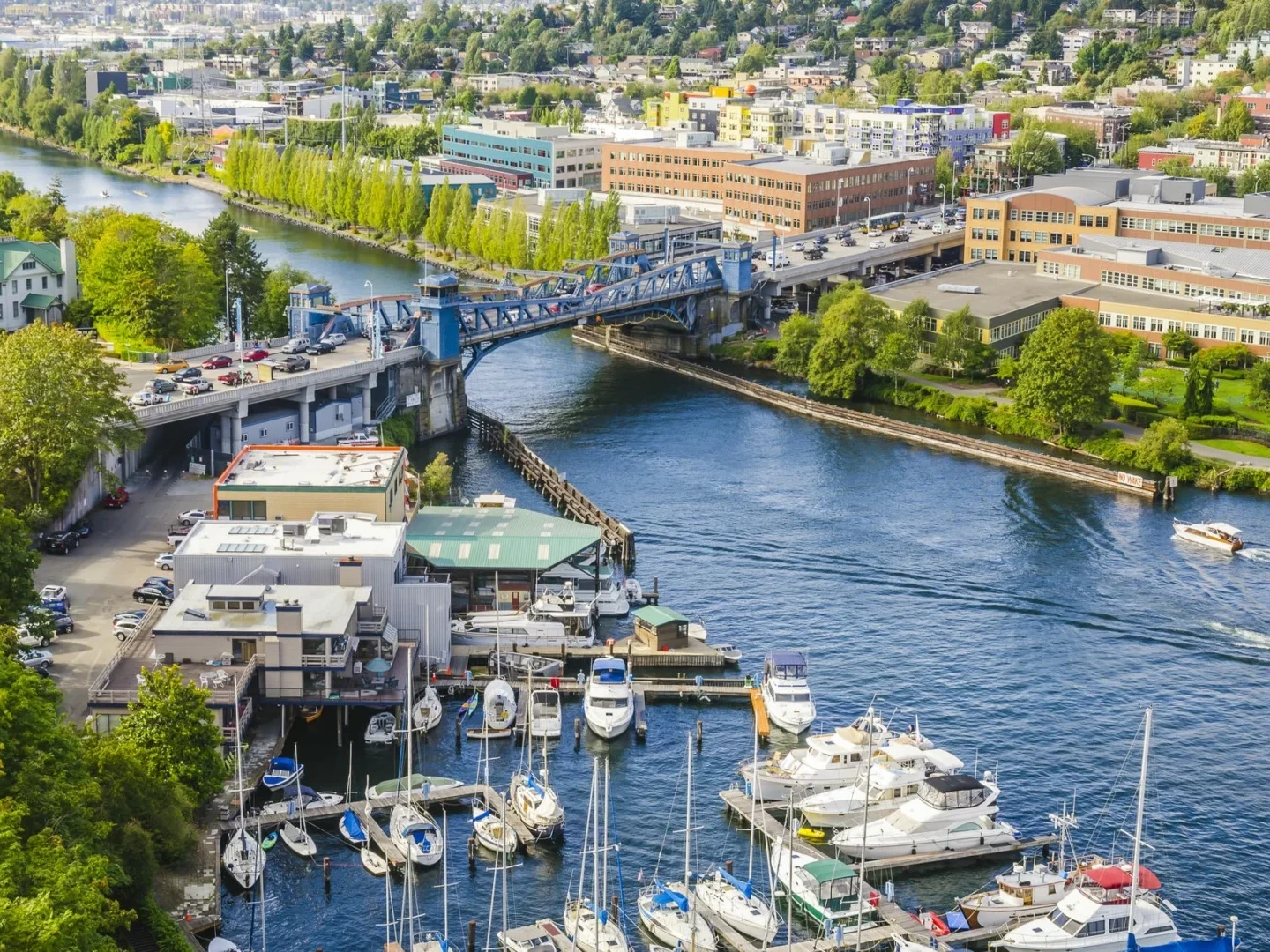 Aerial view of a marina with boats docked, a blue drawbridge over a canal, and surrounding buildings and greenery, with a cityscape in the background.