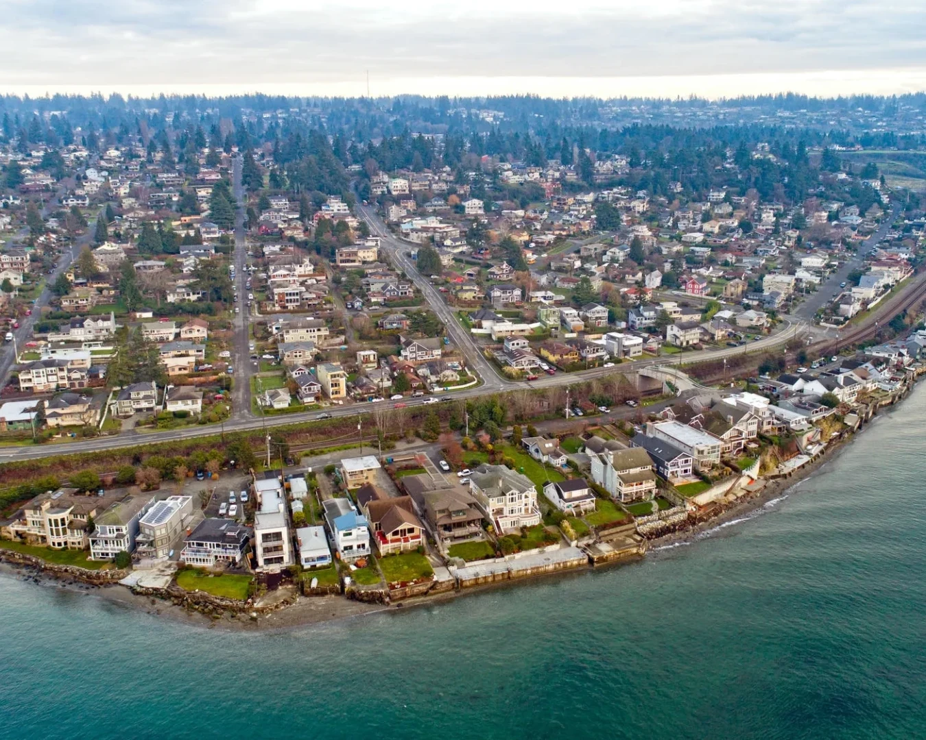 Aerial view of a coastal neighborhood with rows of houses along the shoreline, roads, and lush greenery, with water in the foreground and hills with more houses in the background under a cloudy sky.