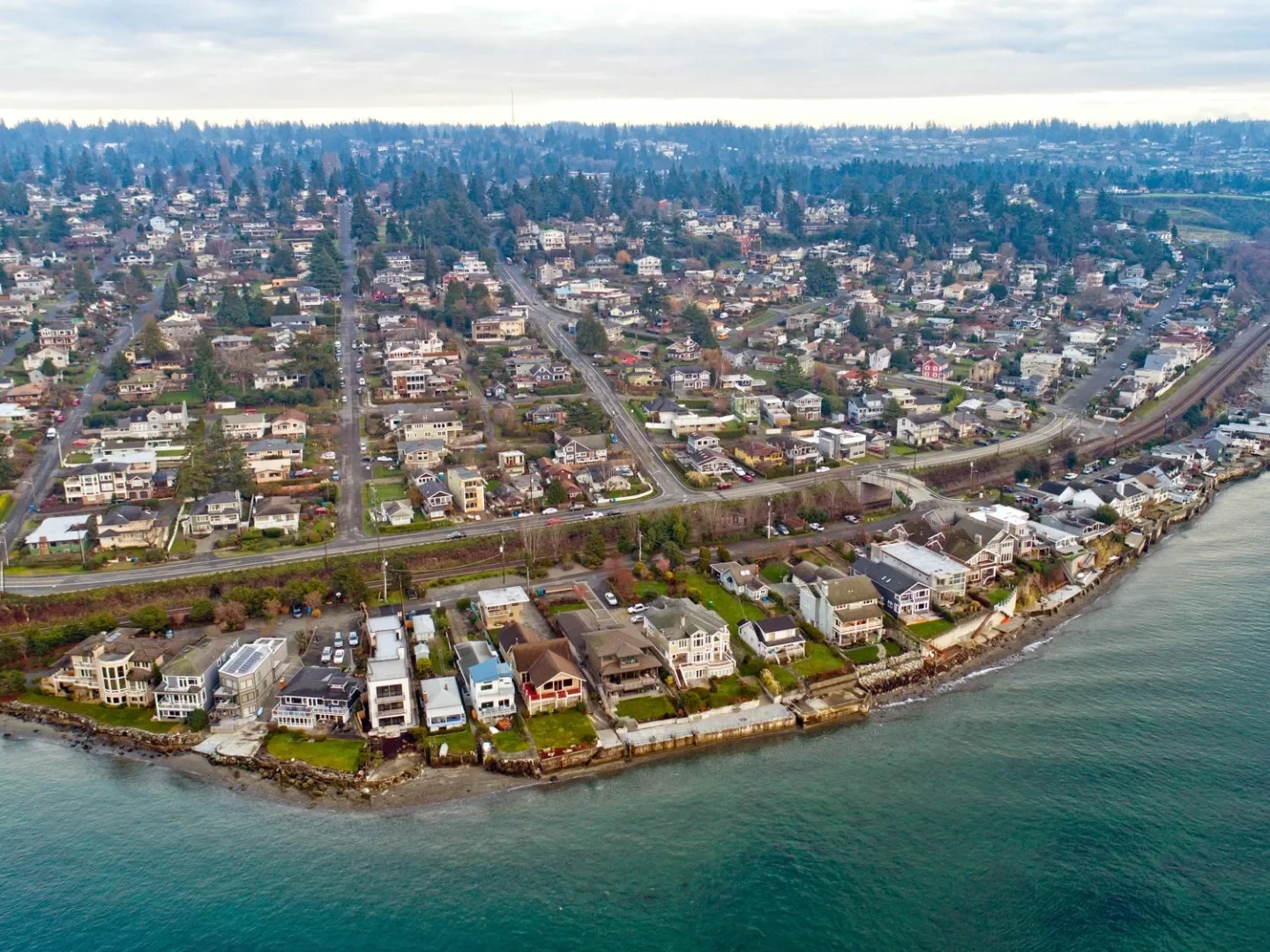 Aerial view of a coastal neighborhood with rows of houses along the shoreline, roads, and lush greenery, with water in the foreground and hills with more houses in the background under a cloudy sky.