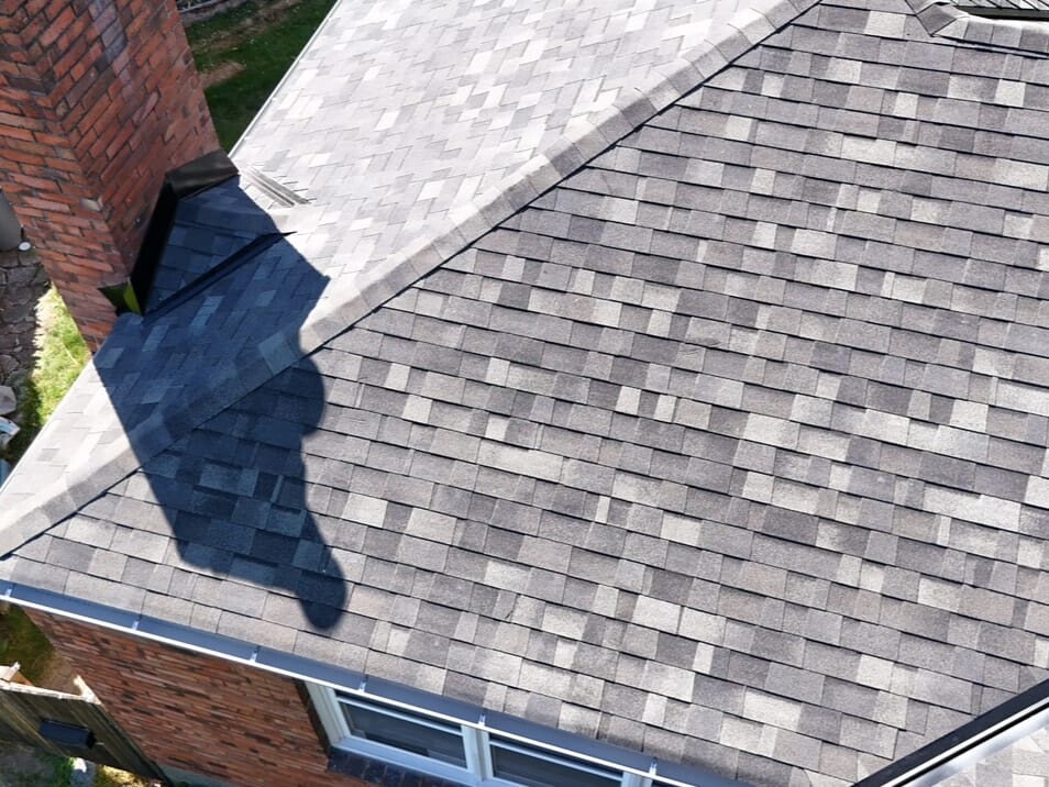 Aerial view of a house roof with gray asphalt shingles, a brick chimney casting a shadow, and part of a green hedge visible along the edge of the property.