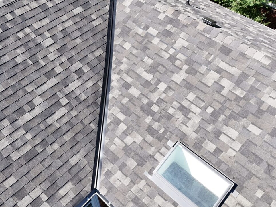Aerial view of a house roof with gray asphalt shingles, showing a roof ridge and a vented skylight window surrounded by patterned shingles. Some greenery and a red brick wall are visible at the top right edge.