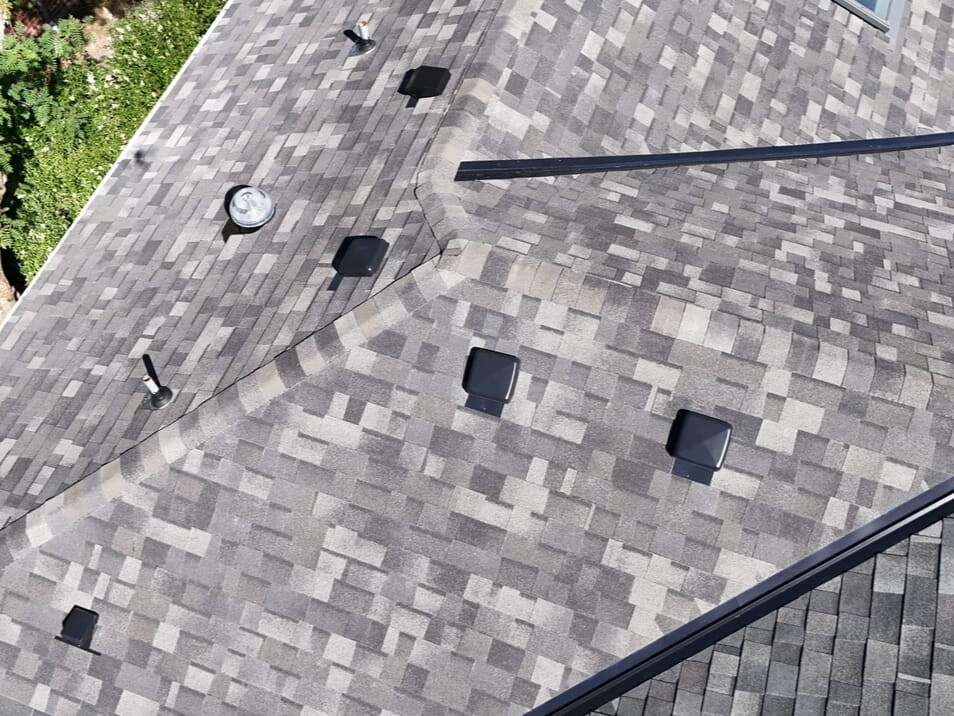 Aerial view of a house roof with gray asphalt shingles, featuring multiple black square vents, a circular metal vent, and nearby green trees.