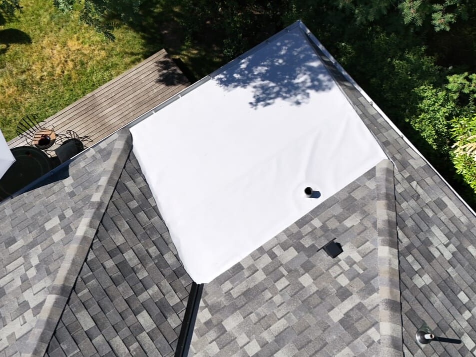 Aerial view of a house roof with gray shingles and a large white tarp covering part of it, surrounded by trees, a wooden deck, and yard furniture visible at the top left.