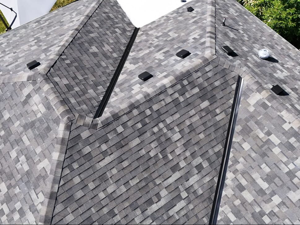 Aerial view of a house roof with gray asphalt shingles, black vents, and metal flashing along the ridges, surrounded by green trees and a patch of grass.