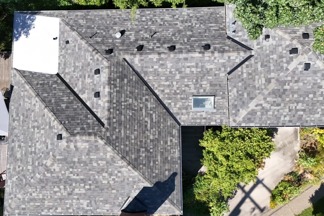 Aerial view of a house roof with gray shingles, multiple angles, one skylight, several vents, and surrounded by green trees and a patio area with an umbrella.