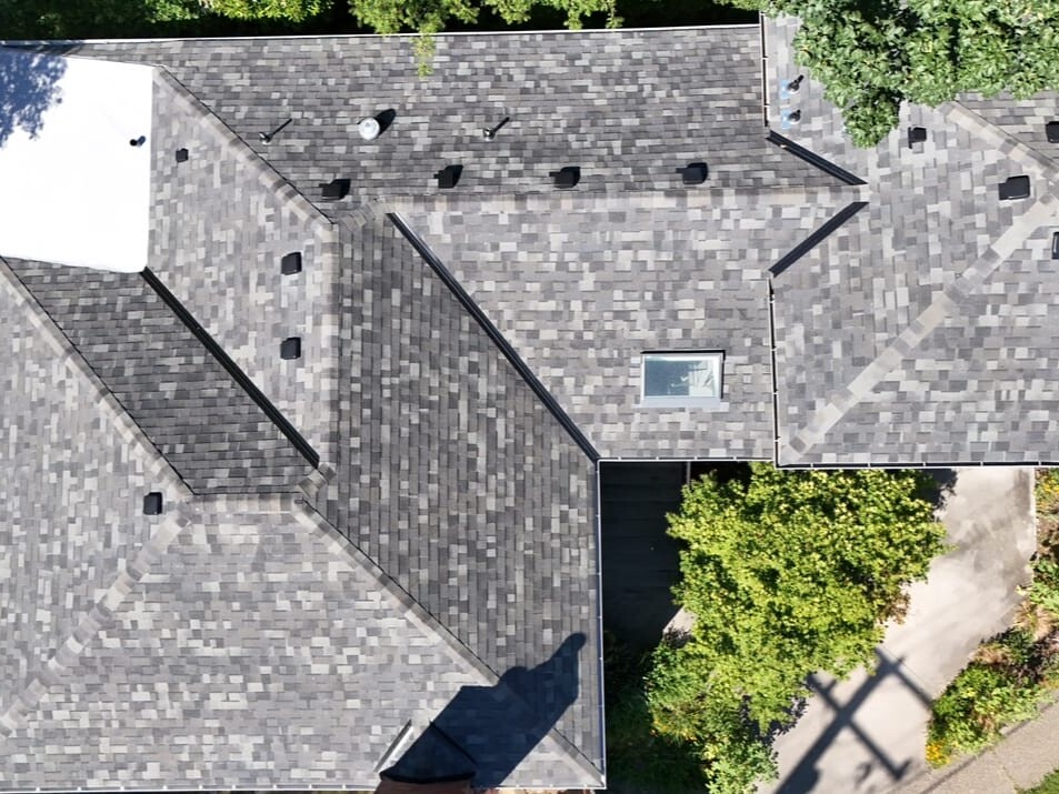Aerial view of a house roof with gray shingles, multiple angles, one skylight, several vents, and surrounded by green trees and a patio area with an umbrella.