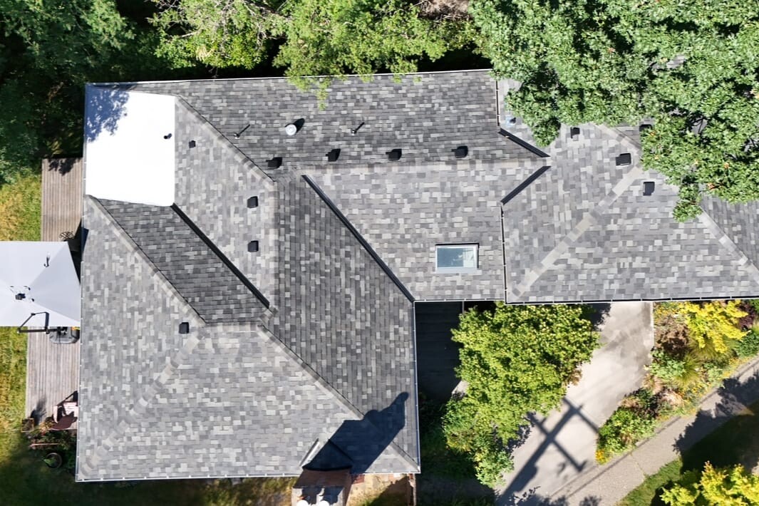 Aerial view of a house with a gray shingled roof surrounded by green trees and landscaping, with patios and outdoor furniture visible on the lawn areas.