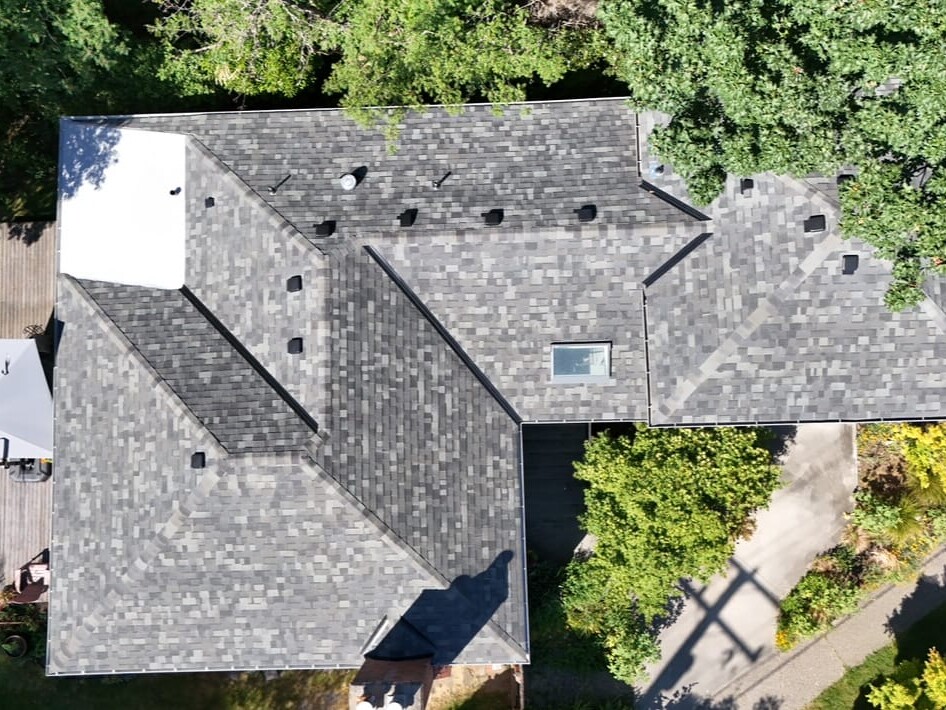 Aerial view of a house with a gray shingled roof surrounded by green trees and landscaping, with patios and outdoor furniture visible on the lawn areas.