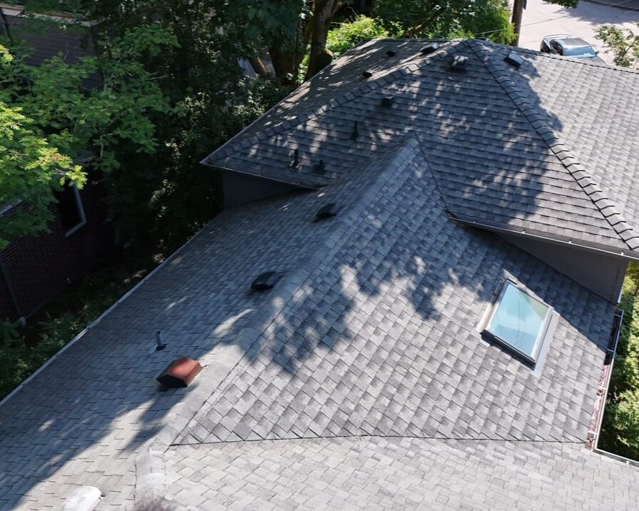 Aerial view of a house roof with gray shingles, several roof vents, and a skylight. Trees cast shadows on the roof, and cars are parked on the street below.