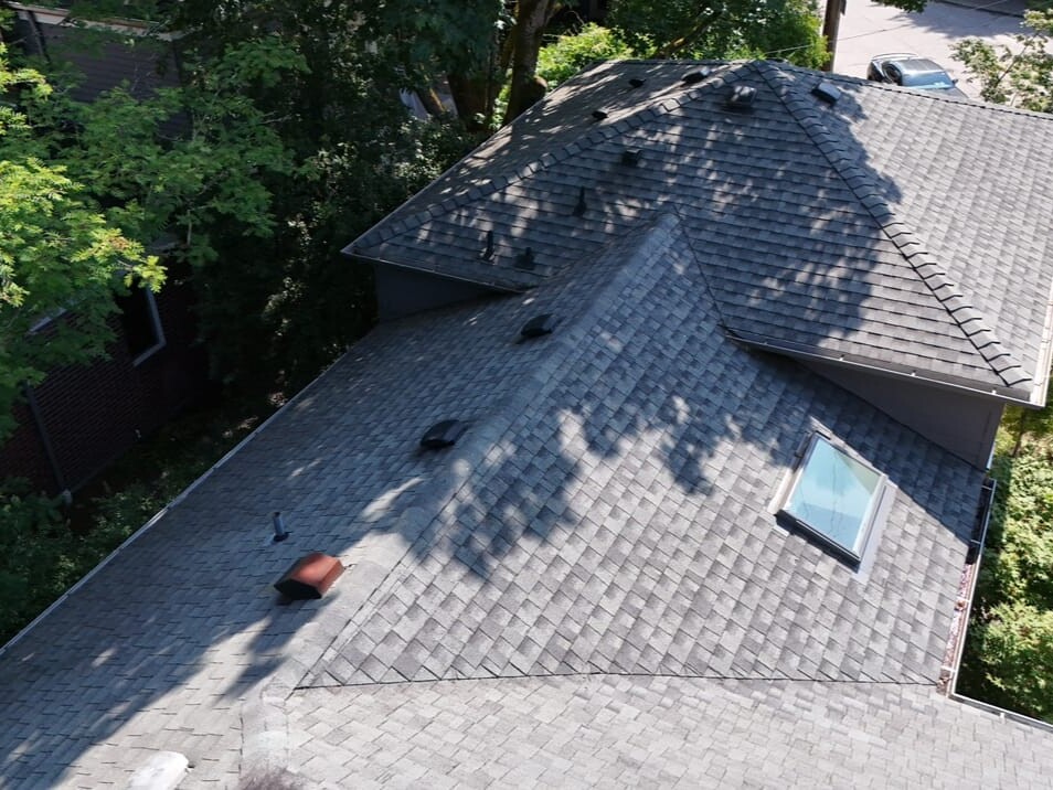 Aerial view of a house roof with gray shingles, several roof vents, and a skylight. Trees cast shadows on the roof, and cars are parked on the street below.