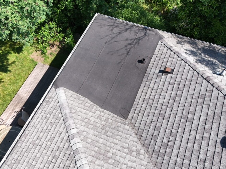 Aerial view of a house roof showing a section with new black roofing material next to older gray shingles, surrounded by trees and a grassy backyard with outdoor furniture.