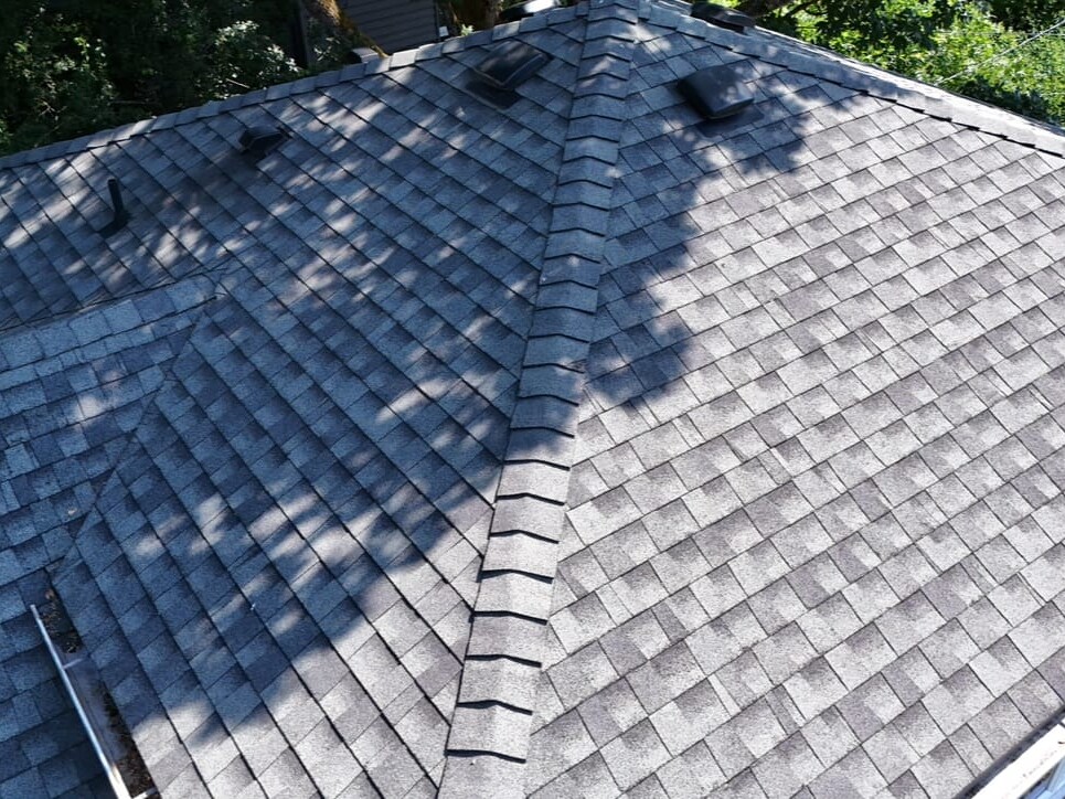 Aerial view of a residential roof with gray asphalt shingles, featuring several roof vents and partial shadows from nearby trees.