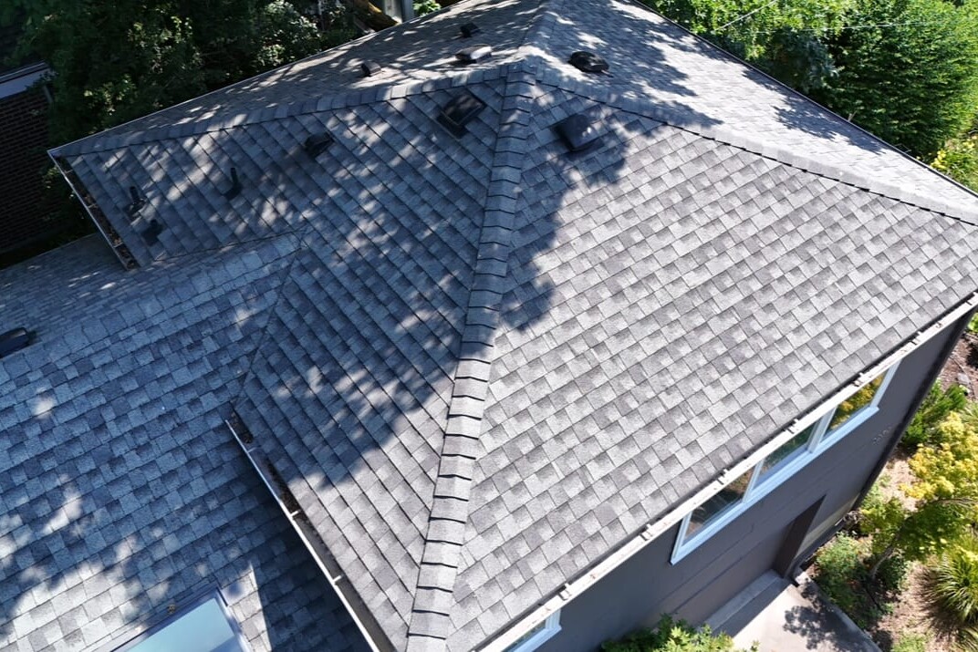Aerial view of a house roof with gray asphalt shingles, multiple vents, and sunlight casting shadows from nearby trees; part of the yard with green bushes and a sidewalk is visible.