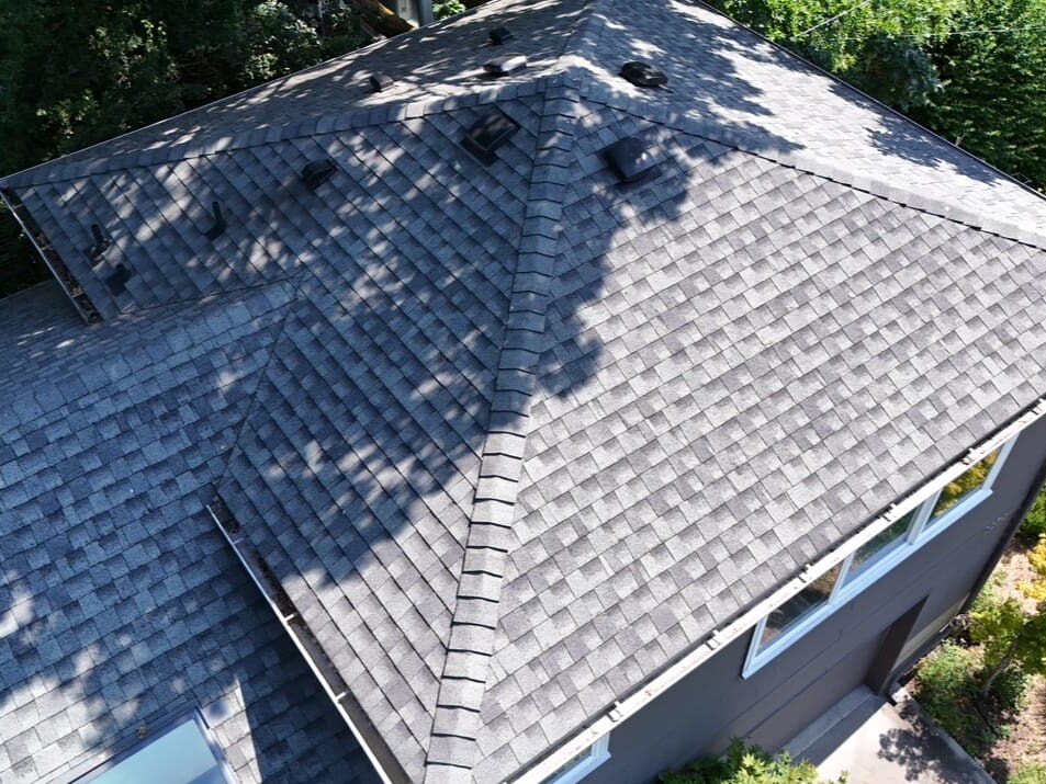 Aerial view of a house roof with gray asphalt shingles, multiple vents, and sunlight casting shadows from nearby trees; part of the yard with green bushes and a sidewalk is visible.