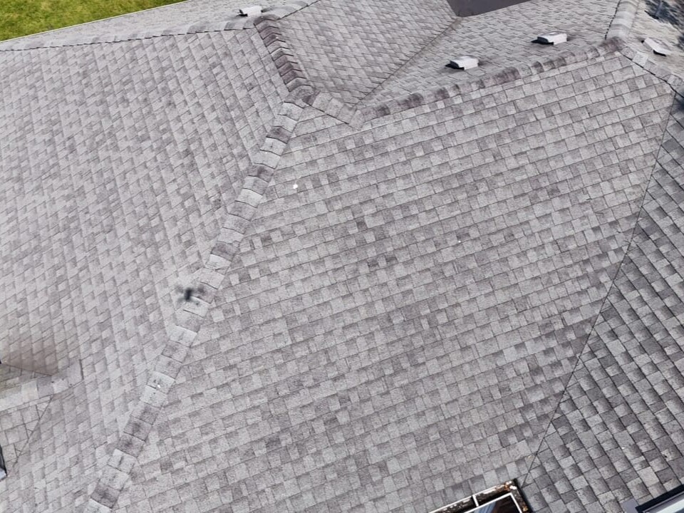 Aerial view of a residential roof with grey asphalt shingles, multiple roof ridges, a brick chimney, roof vents, and a small skylight. Green grass surrounds the house.