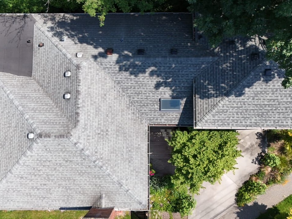 Aerial view of a gray-shingled house roof with a skylight, surrounded by trees, a driveway, garden, and a patio area with outdoor furniture on the left.