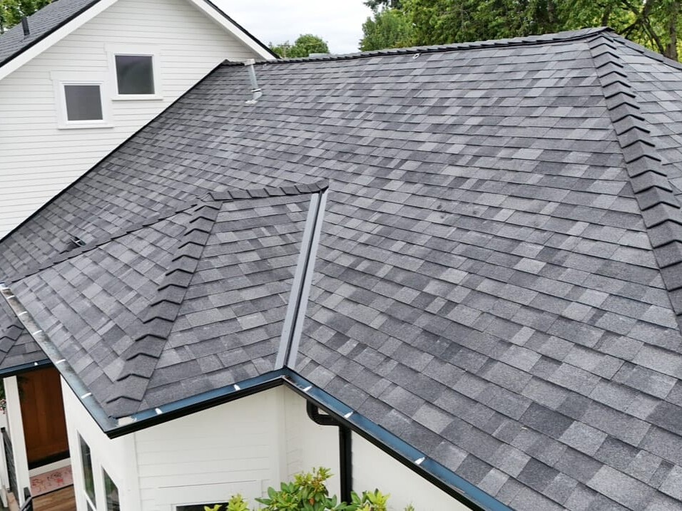 A close-up view of a house’s gray asphalt shingle roof with multiple roof angles and peaks, surrounded by trees and sections of a white exterior.