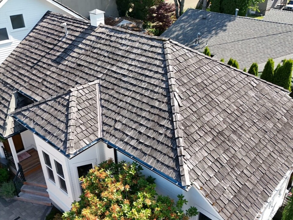 Aerial view of a house with a weathered wooden shingle roof, surrounded by neighboring houses, trees, and shrubs. The roof shows signs of aging and discoloration.