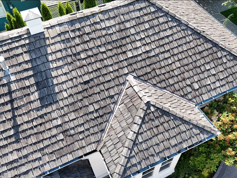 An aerial view of a house with a weathered wooden shingle roof, showing multiple roof angles and a chimney. Green trees and part of a driveway are visible around the house.