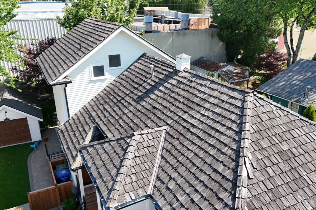 Aerial view of a residential house with dark wooden roof shingles, a detached garage, green lawn, and trees surrounding the property on a sunny day.