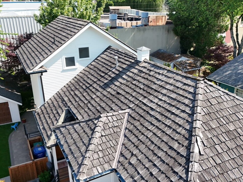 Aerial view of a residential house with dark wooden roof shingles, a detached garage, green lawn, and trees surrounding the property on a sunny day.