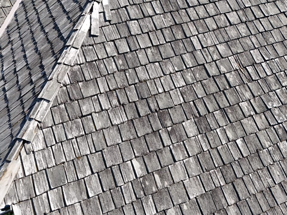 A close-up aerial view of a weathered wooden shingle roof, showing overlapping, grayish shingles with some visible wear and irregular edges. The peak of the roof and part of a green shrub are also visible.