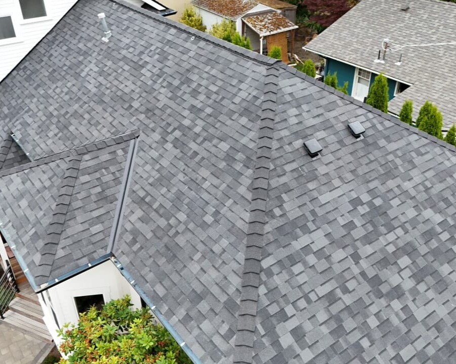 Aerial view of a residential house roof with gray asphalt shingles, vents, and surrounding greenery and other homes visible.