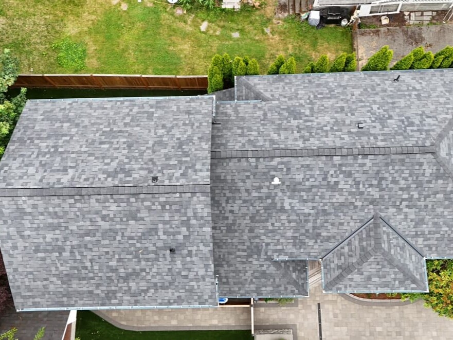 Aerial view of a house with a grey shingled roof, surrounded by a green yard, trees, and a driveway; the image shows the roof’s layout and nearby landscaping.
