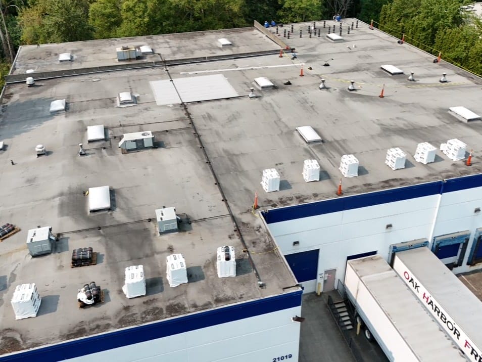 Aerial view of a large flat-roofed industrial building surrounded by trees. The roof is covered with white HVAC units, vents, pipes, and several orange safety cones. Part of the buildings blue and white exterior is visible.