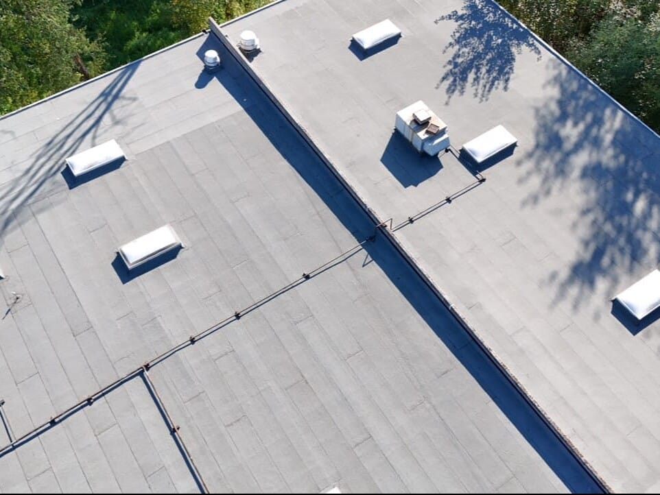 Aerial view of a flat, gray commercial building roof with multiple white vents and skylights, surrounded by green trees and casting shadows on the surface.